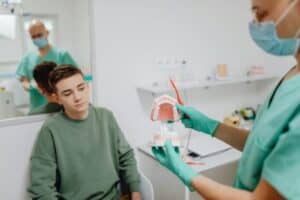 Why Your Gums Bleed When You Brush 3 - Olathe Dentist | Your Olathe Dentist Dentist in green scrubs shows a denture model to a patient seated in a dental chair, while another clinician looks on in the background.