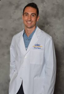 Portrait of a man in a white dental lab coat over a blue checkered shirt, smiling at the camera with UMKC School of Dentistry Tony Raper logo on the coat