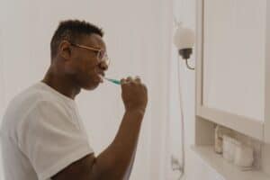 Family Dentist in Olathe - Olathe Dentist | Your Olathe Dentist Man wearing glasses brushing his teeth with a toothpaste-coated toothbrush in a bathroom.
