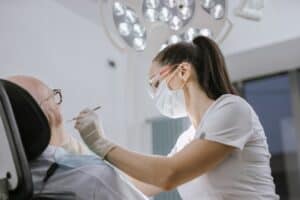 Olathe Dentist Examining Patient - Olathe Dentist | Your Olathe Dentist Female dentist in a mask and gloves treats an elderly patient in a bright dental clinic.